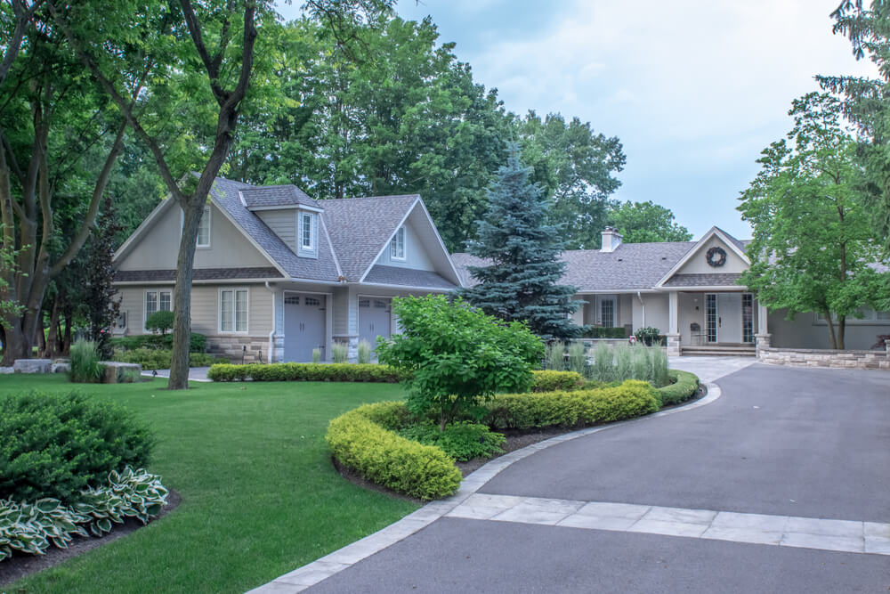 A Beautiful Large Custom Bungalow Home With a Grey Brick and Stone Exterior and a 3 Car Garage, on a Quite Town Street With a Winding Circular Paved Driveway and Lush Trees, Gardens and Lawn.