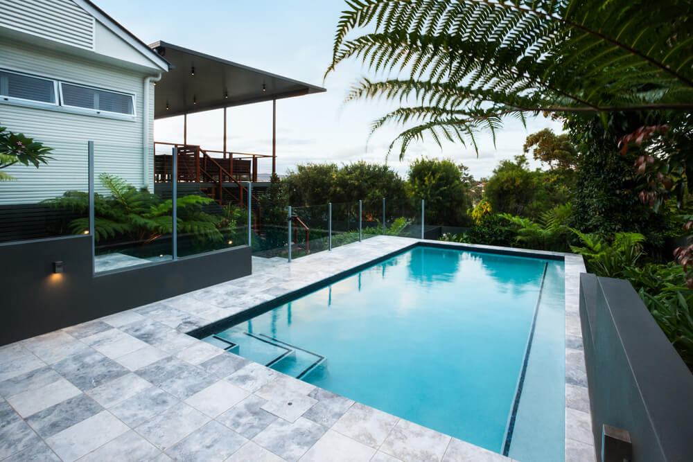 Blue Water Swimming Pool With Floor Tiles Under Green Trees Close Up With a Garden Including a Bunch of Green Leaves