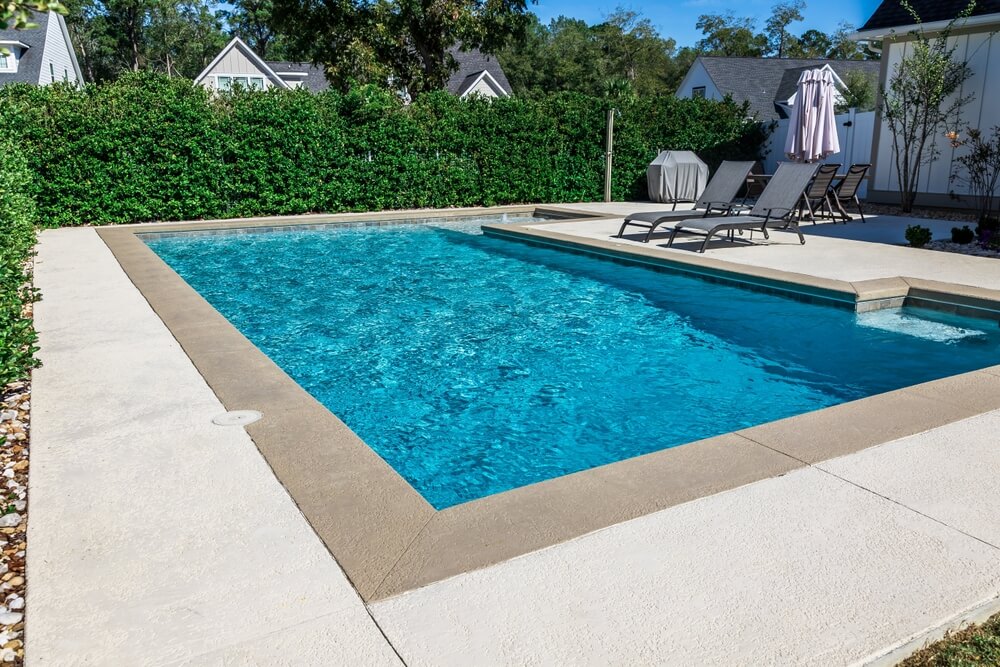 A Rectangular New Swimming Pool With Tan Concrete Edges In The Fenced Backyard Of A New Construction House With Privacy Hedge